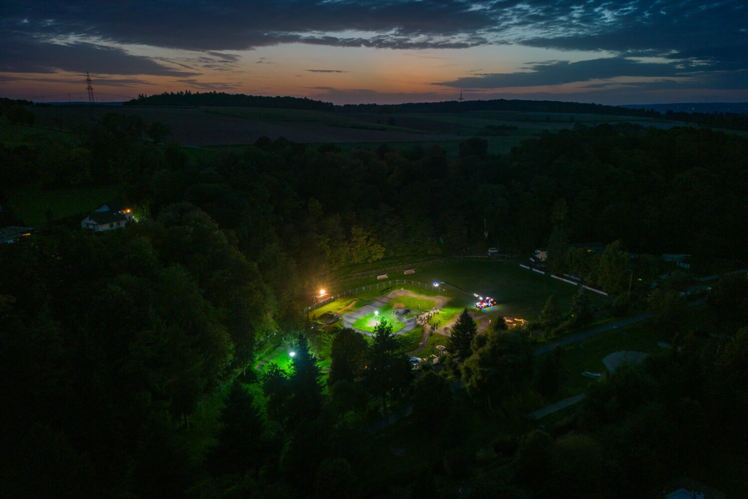 Businessfotografie für Unternehmen: Drohnenaufnahme eines geschwungenen Biketracks auf einer Grünfläche, mit asphaltierten Wegen, einzelnen Personen und einem Einsatzfahrzeug der Feuerwehr, das den Biketrack in der Dämemrung beleuchtet am Rand.