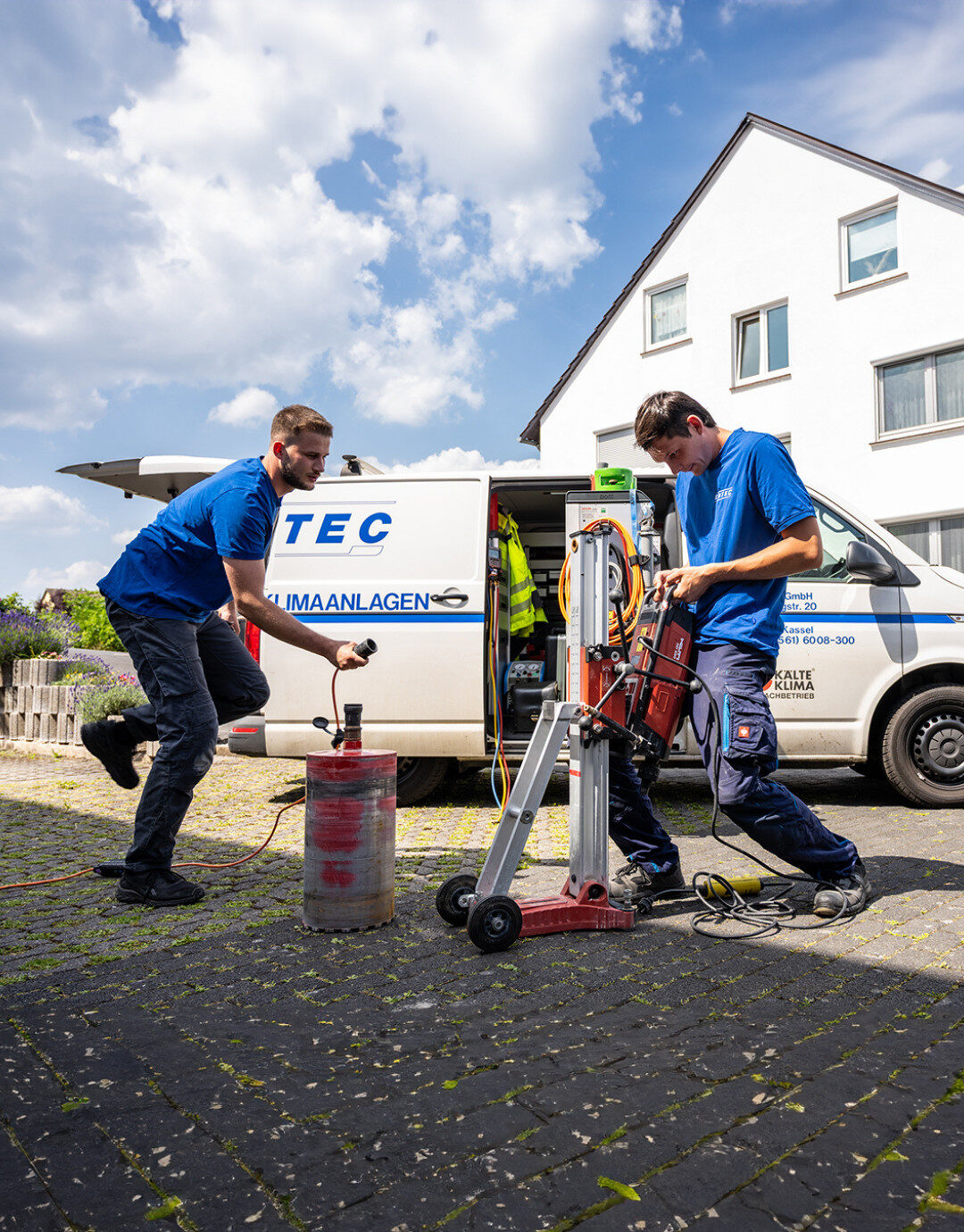 Zwei Servicetechniker führen vor einem Firmenfahrzeug Bohrarbeiten im Außenbereich durch und bereiten eine technische Installation vor.