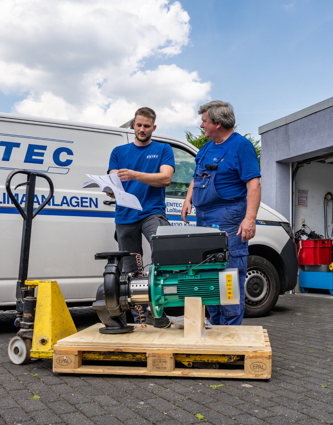 Zwei Mitarbeiter stehen vor einem Servicefahrzeug und prüfen anhand von Unterlagen eine technische Anlage auf einer Palette.