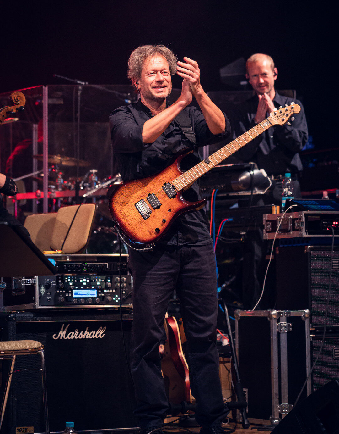 Businessfotografie für Unternehmen: Gitarrist eines Philharmonieorchesters steht auf der Bühne und applaudiert dem Publikum, die E-Gitarre hängt umgehängt vor dem Körper, im Hintergrund sind weitere Orchestermitglieder, Instrumente und Bühnentechnik bei Konzertbeleuchtung zu sehen.