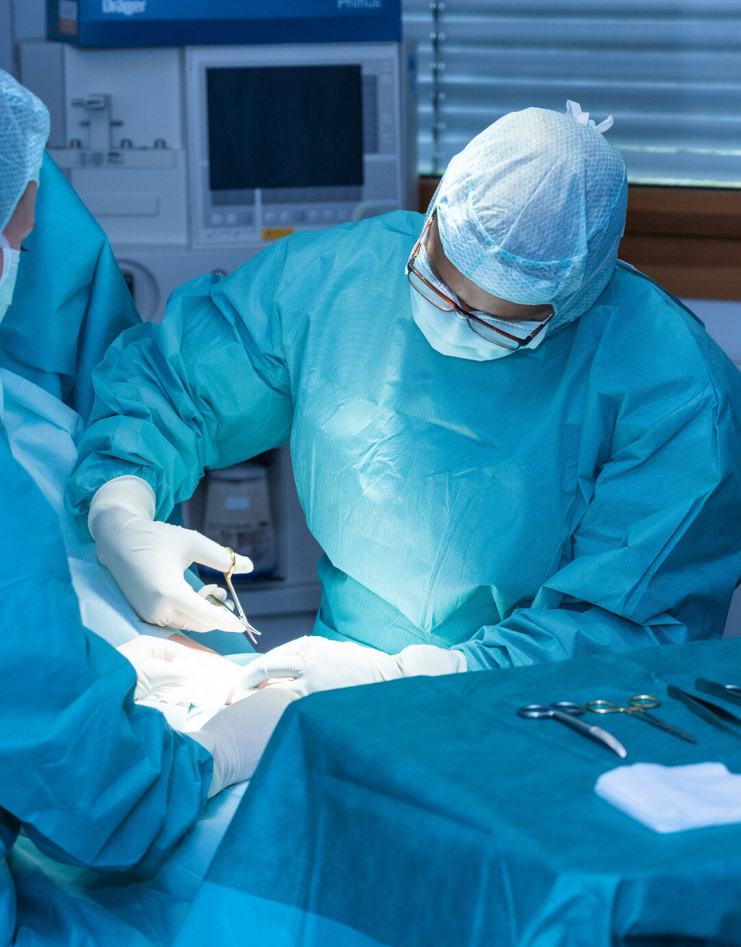 Businessfotografie für Unternehmen: Zwei medizinische Fachkräfte in steriler Schutzkleidung führen gemeinsam einen chirurgischen Eingriff in einem Operationssaal durch; OP-Instrumente liegen auf einem abgedeckten Tisch, medizinische Geräte sind im Hintergrund sichtbar.