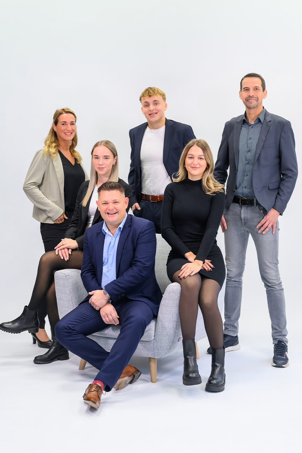 Businessfotografie für Unternehmen: Gruppenporträt eines Teams aus sieben Personen in Businesskleidung, teils stehend, teils sitzend auf einem Sessel, vor hellem Studiohintergrund, freundliche und professionelle Darstellung des gesamten Teams.