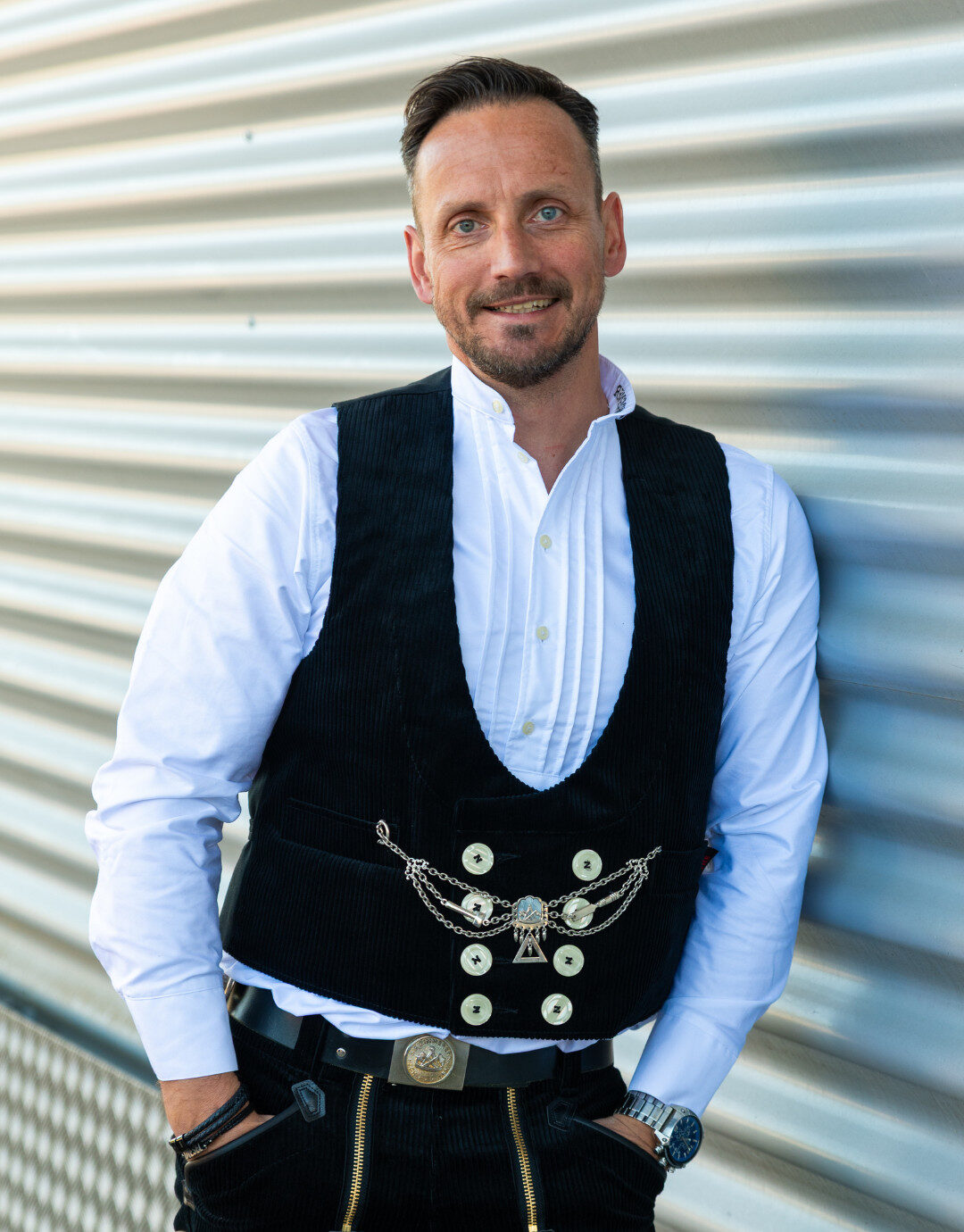 Businessfotografie für Unternehmen: Porträt eines Mannes in traditioneller Arbeitskleidung mit Weste und weißem Hemd, der entspannt an einer metallischen Wand lehnt und in die Kamera lächelt, Aufnahme im Außenbereich mit klarer, natürlicher Lichtführung.