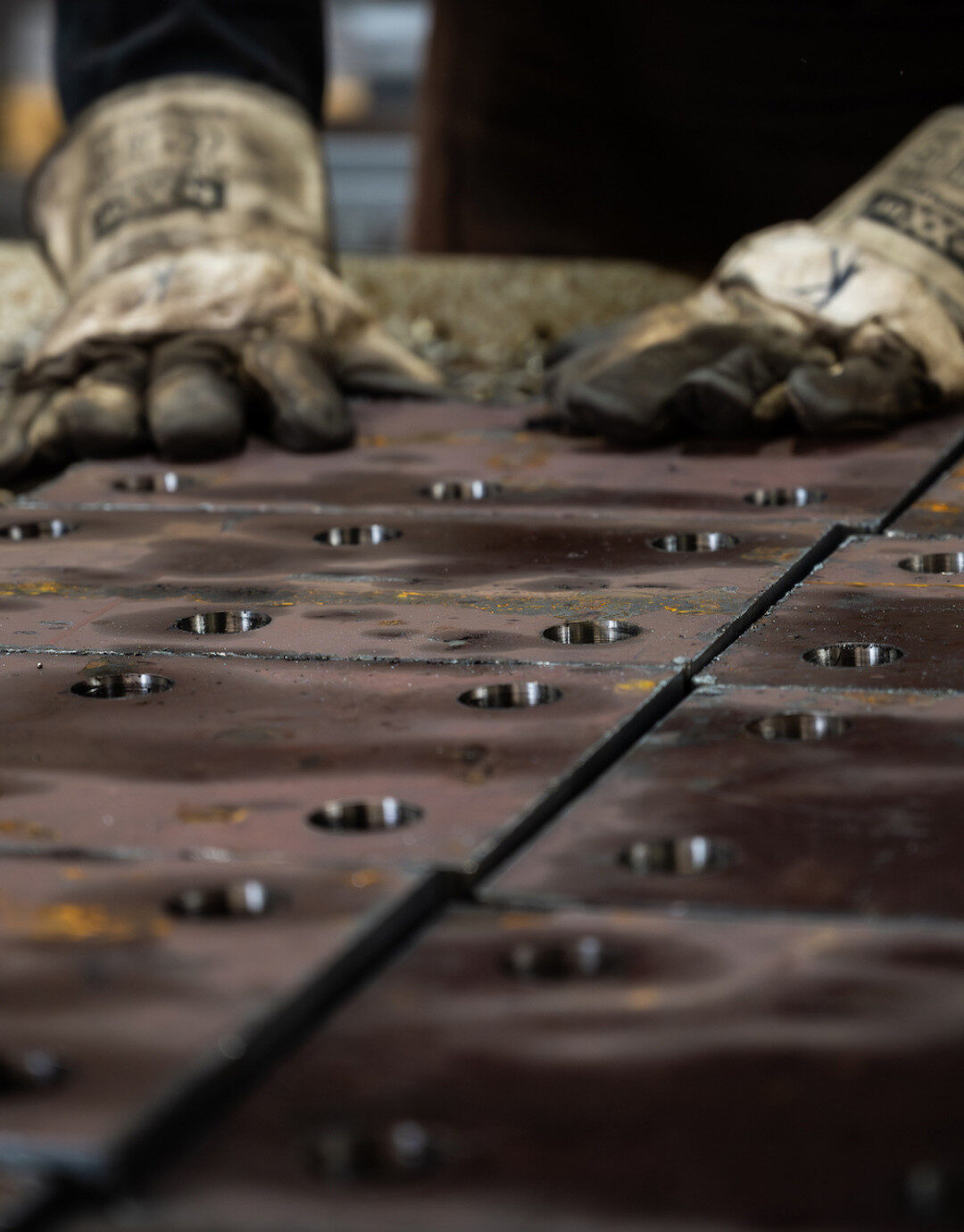 Businessfotografie für Unternehmen: Detailaufnahme eines industriellen Arbeitsprozesses: Zwei Arbeitshandschuhe liegen auf mehreren metallischen Bauteilen mit Bohrungen, Oberfläche zeigt Gebrauchsspuren und Bearbeitung, Fokus auf Material und Struktur, Hintergrund unscharf.