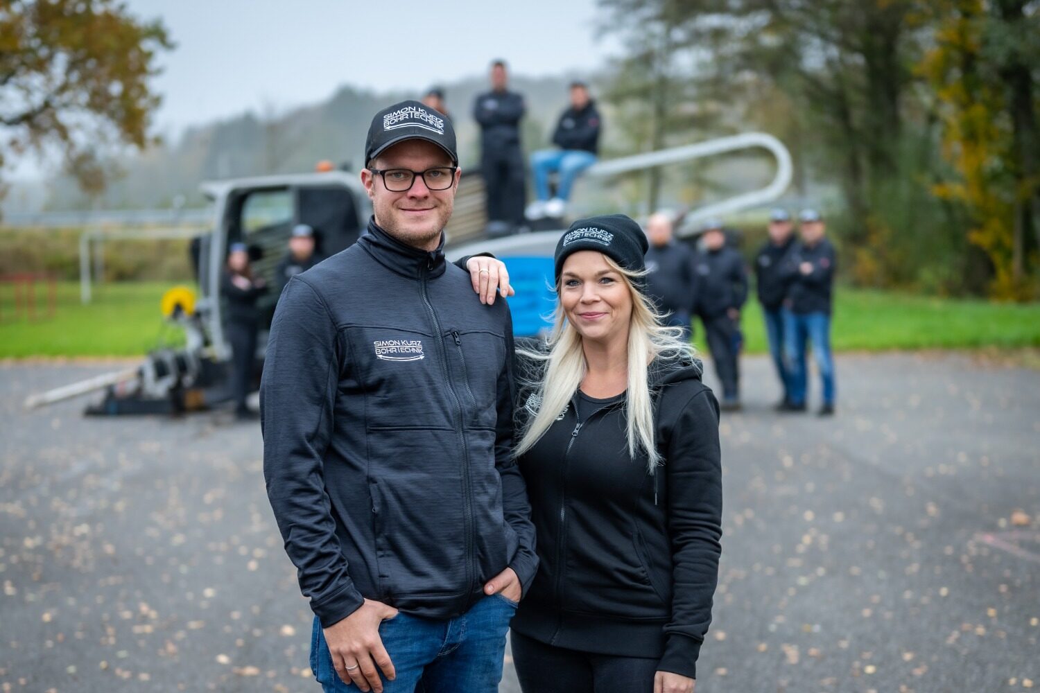 Zwei Mitarbeitende eines Handwerksbetriebs stehen vor ihrem Team und einem Arbeitsfahrzeug auf einem Betriebsgelände.