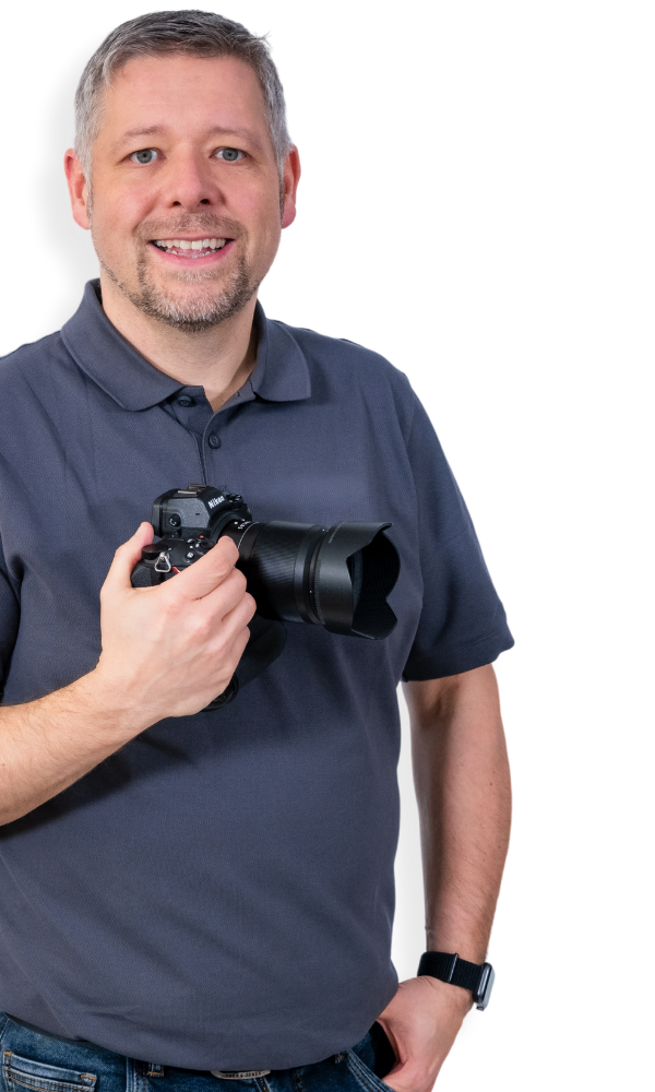 Porträt von Martin Diebel vor freigestelltem Hintergrund, der eine Kamera in der Hand hält und ruhig in die Kamera blickt, Darstellung einer persönlichen und professionellen Arbeitsweise