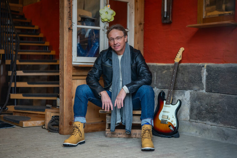 Mann mit Brille sitzt vor einer roten Hauswand neben einer Gitarre